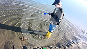 Boy in yellow boots, blue cap, playing in ocean with a live crab