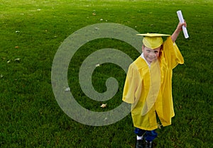 Boy in yellow preschool graduation gown, holding up his diploma