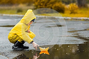Boy in yellow waterproof cloak and rubber boots playing with paper handmade boat toy in a puddle outdoors in the rain in autumn