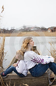 Boy and a young woman in embroidered national clothes are sitting back to back on the shore of the lake