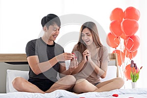 The boyfriend surprised his girlfriend with a ring contained in a red ring box on the bed in the bedroom decorated with red heart.