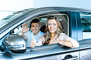 Boyfriend waving a hand, girlfriend showing thumb up sitting in a new car in showroom