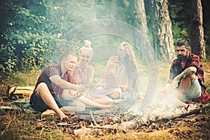 Boyfriends and girlfriends enjoy camping food. Friends have picnic at bonfire in forest. Men and women roast sausages on