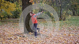 Boys run around the trunk of a large tree and toss the autumn yellow leaves up.