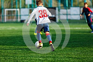 Boys in white and blue sportswear plays football on field, dribbles ball. Young soccer players with ball on green grass
