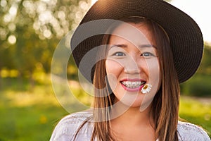 Braces on the teeth, and in the mouth a chamomile flower, a girl of Asian appearance in a white T-shirt and a dark hat