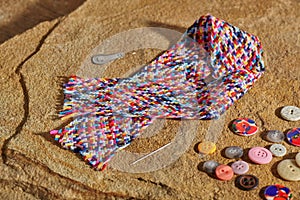 A braid of multi colored sewing threads, needle, needle threader, buttons lying on a stone