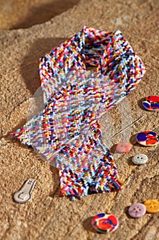 A braid of multi colored sewing threads, needle, needle threader, buttons lying on a stone