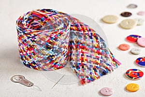 A braid of multi colored sewing threads, needle, needle threader, buttons lying on a white surface