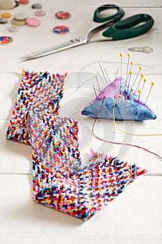 A braid of multi colored sewing threads, pincushion, scissors, needle threader, buttons lying on a white surface