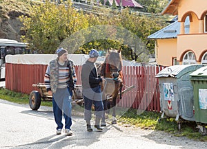 Two men lead by the bridle of a horse harnessed to a wagon in a suburb of Bran city in Romania