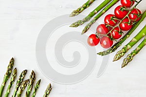 A branch of fresh cherry tomatoes and some asparagus stems in two opposite corners of the off-white rustic table.