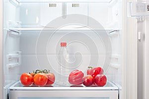 Branch of red tomatoes, two red peppers, two two-colored orange and red peaches and a bottle of water on shelf of open empty