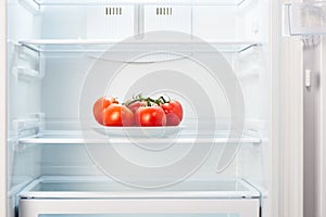 Branch of red tomatoes on white plate in open empty refrigerator