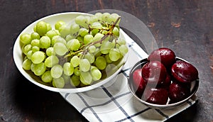 Branch of ripe green grape on plate with water drops and plums in bowl. Juicy fruits on wooden background, closeup