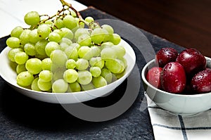 Branch of ripe green grape on plate with water drops and plums in bowl. Juicy fruits on wooden background, closeup