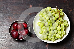 Branch of ripe green grape on plate with water drops and plums in bowl. Juicy fruits on wooden background, closeup