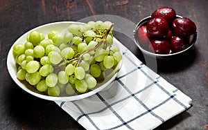 Branch of ripe green grape on plate with water drops and plums in bowl. Juicy fruits on wooden background, closeup