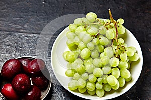 Branch of ripe green grape on plate with water drops and plums in bowl. Juicy fruits on wooden background, closeup