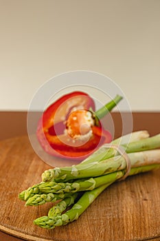 branches of fresh green asparagus on a wooden board, half of a red big pepper in the background. brown and light beige