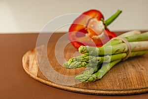 branches of fresh green asparagus on a wooden board, half of a red big pepper in the background. brown and light beige