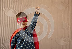 Brave cheerful boy in superhero cape and mask standing with hand on waist on beige background