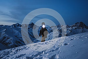 Brave explorer with headlamp commits a night climb to a steep snowy mountain. Wearing ski wear and backpack. Snowboar