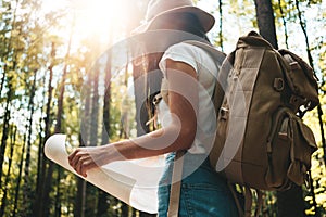 Brave hipster girl traveling alone and looking around in forest on outdoors wearing treveler backpack and hold location map in han