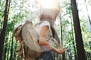 Brave hipster woman wearing backpack and hat traveling alone among trees in forest on outdoors