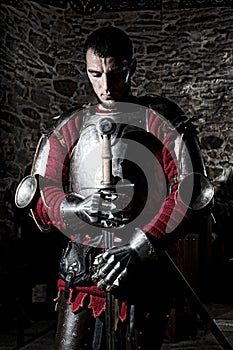 Brave Knight Standing With Head Bowed in Prayer and Holding Metal Sword Against Stone Wall photo