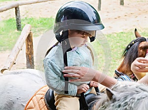 A brave little girl in a helmet rides a little pony in an amusement park. A happy, pretty little girl in a riding helmet sits on a