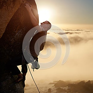 Brave male climber blinded by sunlight on high natural rock wall over gray clouds. Climbing with rope and safety harness