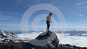 Brave mountaineer in a helmet with trekking poles stands on a large rock above the clouds.