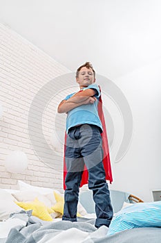 Brave proud little boy in a superhero costume stands on the bed, imagining flying