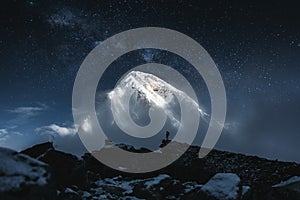 Brave solo mountain traveler stand in front of huge snowy mountain and starry sky. Professional tourist on mountains summit