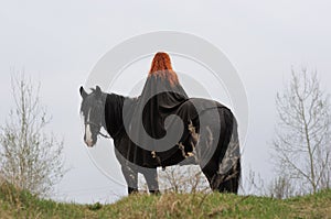 Brave woman with red hair in black cloak on friesian horse