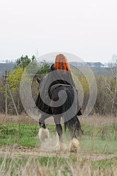 Brave woman with red hair in black cloak on friesian horse