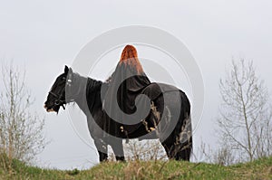 Brave woman with red hair in black cloak on friesian horse