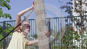 Brave young blonde girl play on playground outdoors, trying to climb to wooden log holding on rope. Close-up. Metal