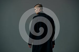 Brave young man dressed in black coat detective or criminal on gray studio background, back view