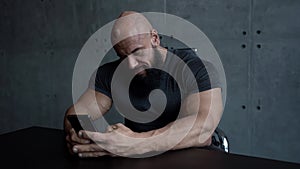 Brawny bald man is sitting at table at home and using smartphone, playing games or checking email