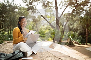 Brazilian lady student writing something in notebook, sitting with laptop in park and studying outdoors, copy space