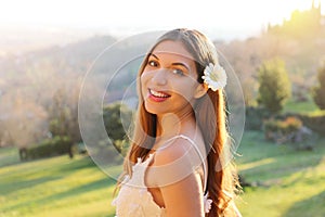 Brazilian smiling girl with white flower on ear and natural background looks at camera in spring time on sunset