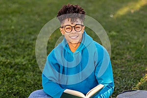 Brazilian student guy wearing glasses happily holding book in park