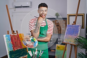 Brazilian young man holding painter palette at artist studio winking looking at the camera with sexy expression, cheerful and