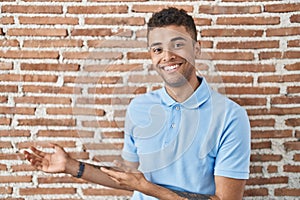 Brazilian young man standing over brick wall inviting to enter smiling natural with open hand