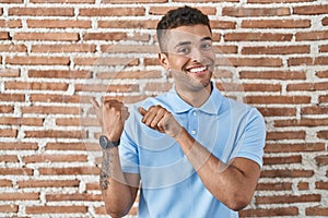 Brazilian young man standing over brick wall pointing to the back behind with hand and thumbs up, smiling confident