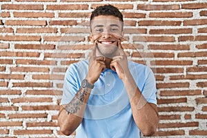 Brazilian young man standing over brick wall smiling with open mouth, fingers pointing and forcing cheerful smile
