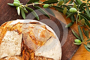 Bread loaf on a wooden board with green olives on branch with leaves over a used oak wood background