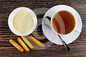 Bread sticks, bowl with condensed milk, spoon, tea in cup on saucer on table. Top view